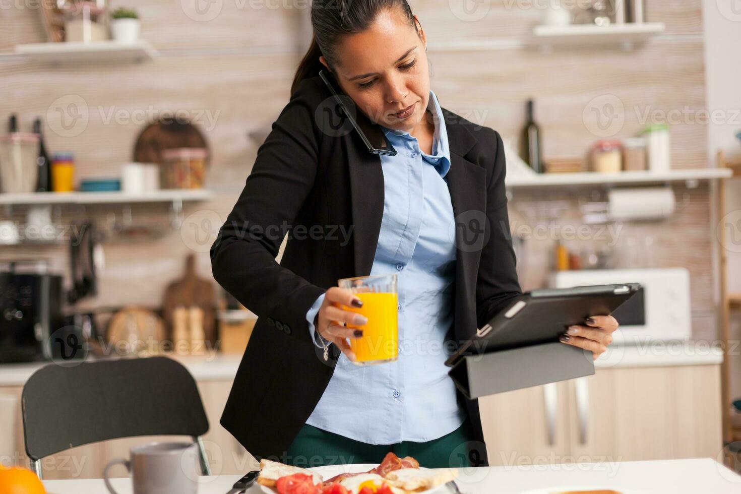 Busy freelancer talking on the phone at breakfast. Concentrated business woman in the morning multitasking in the kitchen before going to the office, stressful way of life, career and goals to meet photo