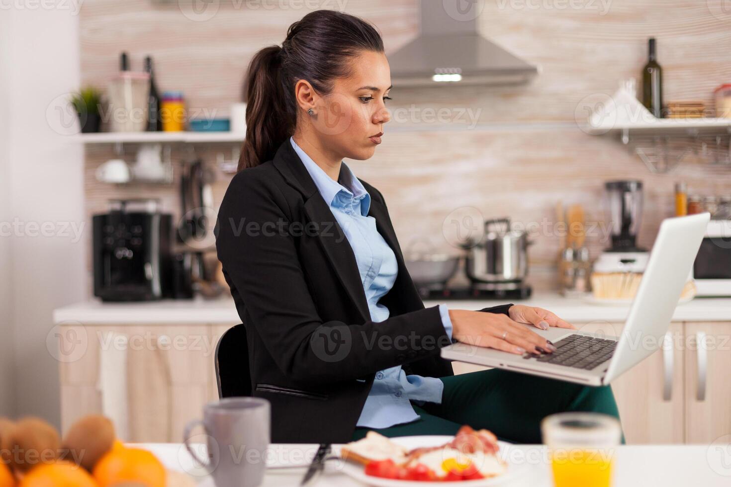 Free agent working at laptop in the morning eating the breakfast. Young freelancer in the kitchen talking on a video call with her colleagues from the office, using modern internet technology photo