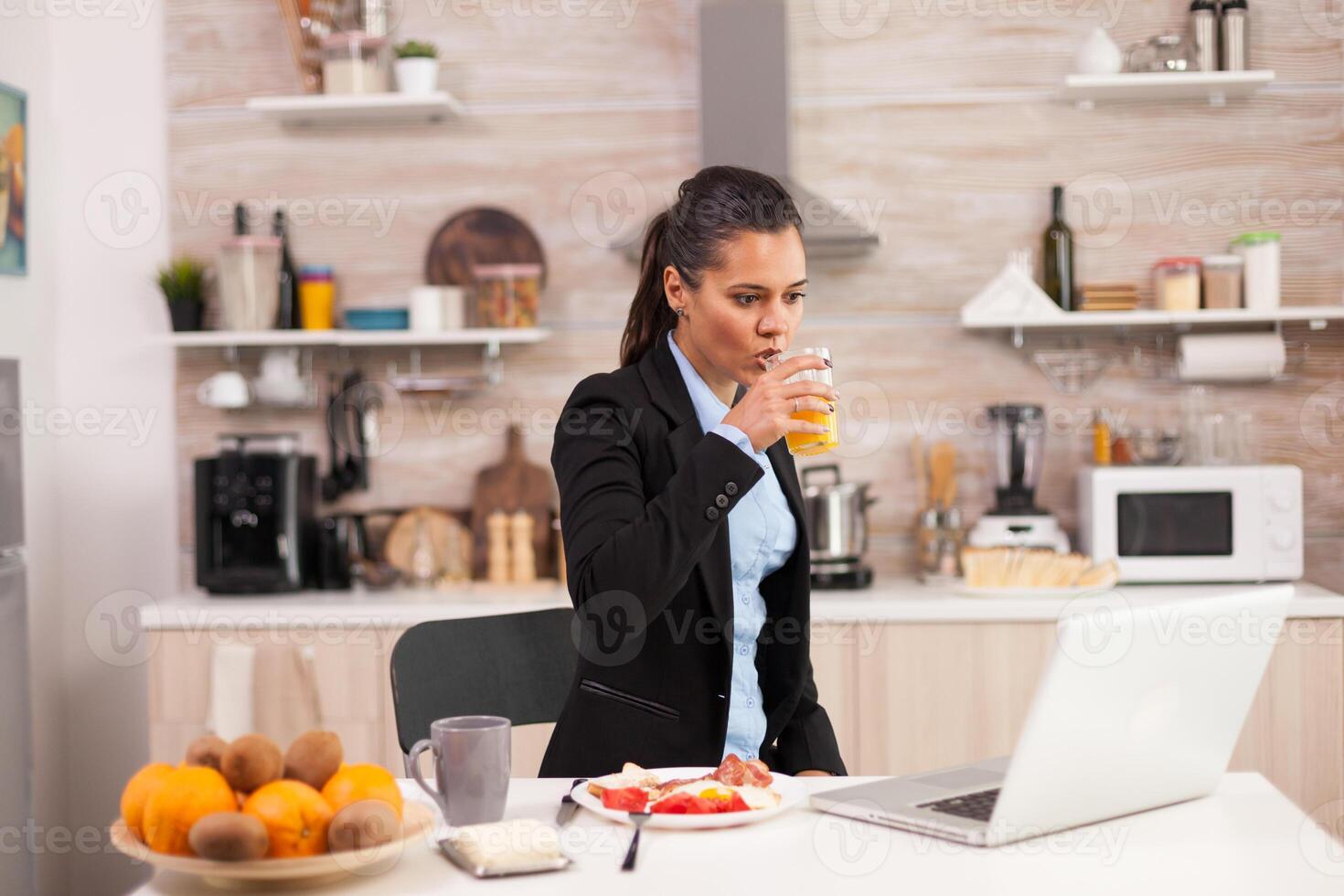 Young business woman enjoying the breakfast and looking at laptop. Business woman reading the last news online before going to work, using modern technology in the kitchen while eating a healthy meal photo