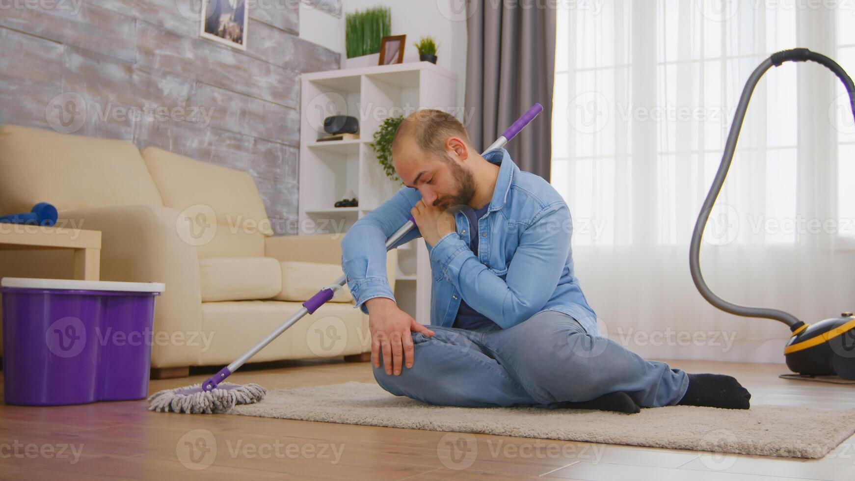 Tired man sitting on cozy carpet after cleaning the room floor with mop and detergent. photo
