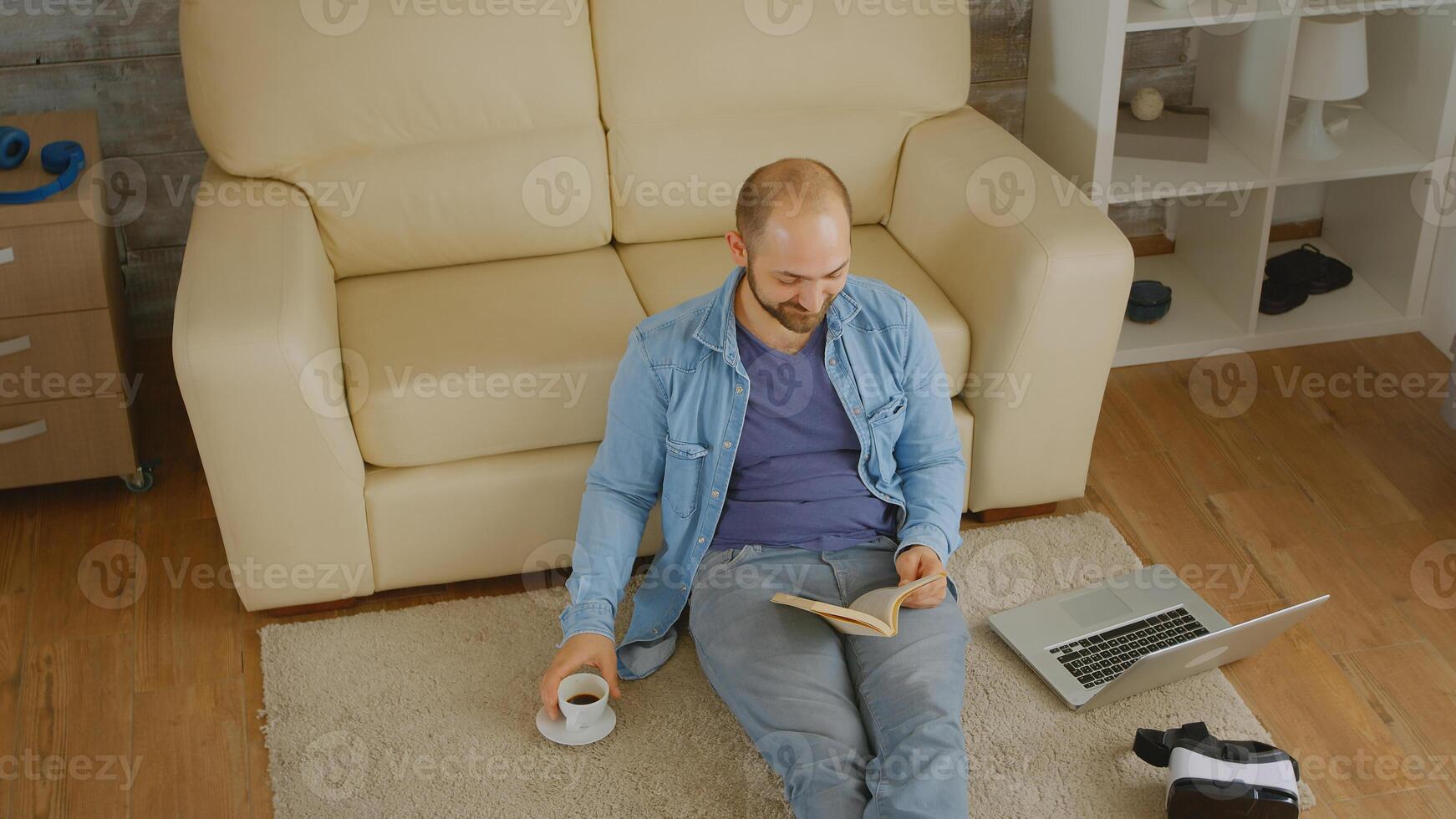 Man in denim shirt laughing while reading a book and drinking some coffee on the floor photo