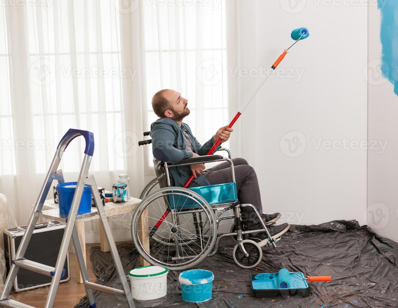 Invalid man painting wall while sitting in wheelchair. Handicapped, disabled ill and immobilizes man helping with apartment redecoration and home construction while renovating and improving, photo