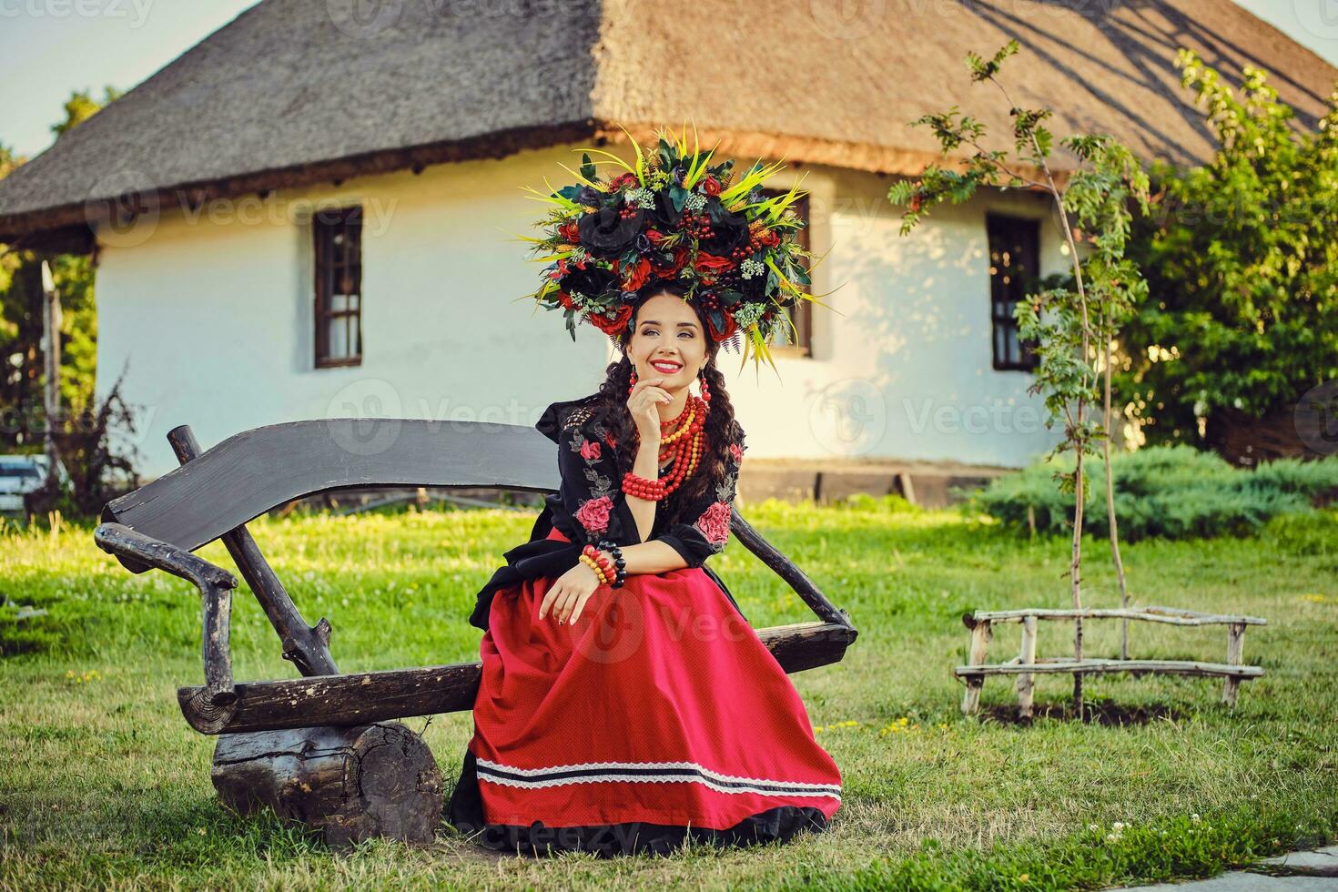 Brunette girl in a white ukrainian authentic national costume and a wreath of flowers is posing sitting on a wooden bench against a white hut. photo