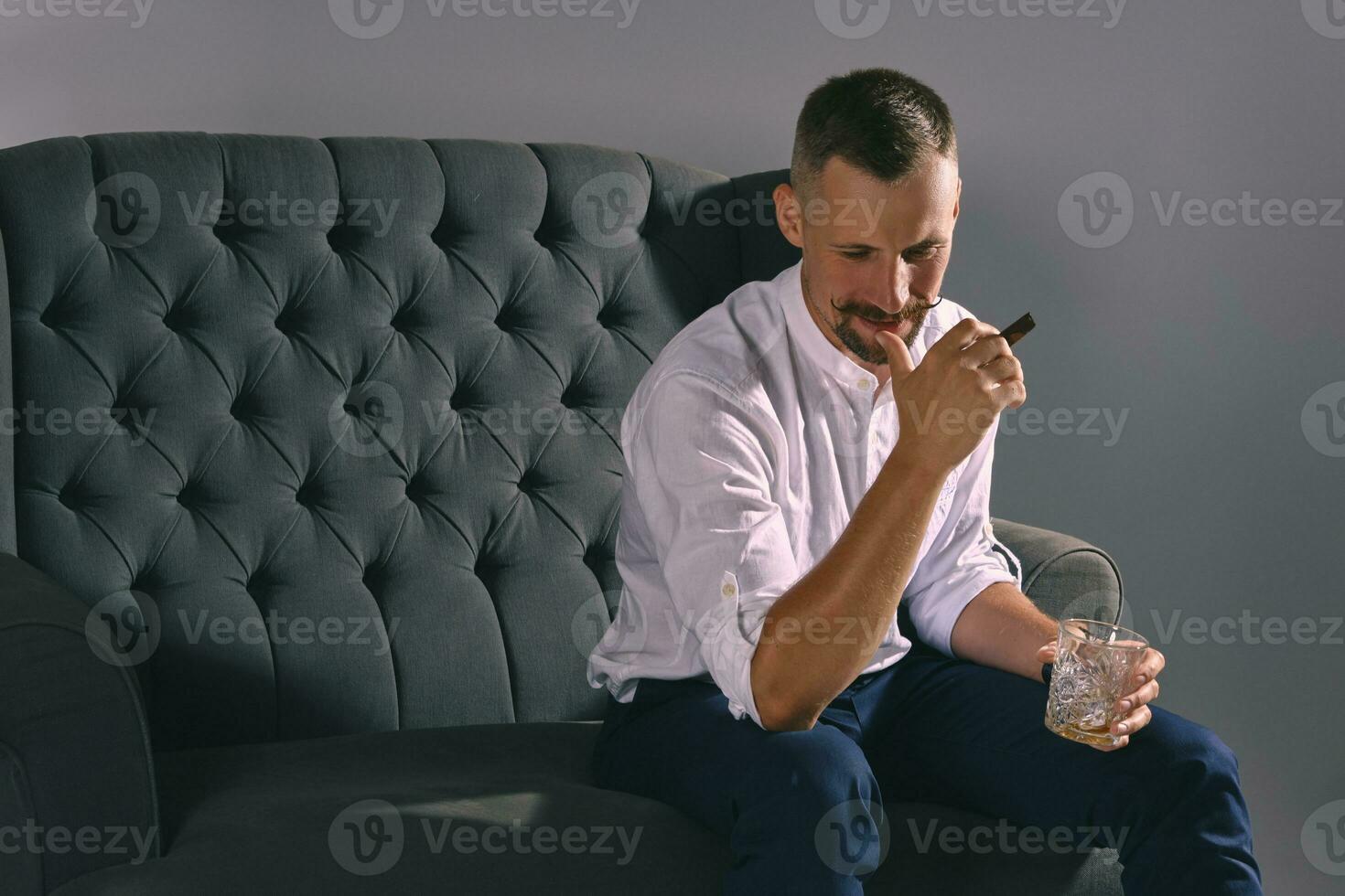 Man with stylish mustache, dressed in white shirt, blue trousers is sitting on dark sofa, holding glass of whiskey, cigar. Grey background, close-up. photo
