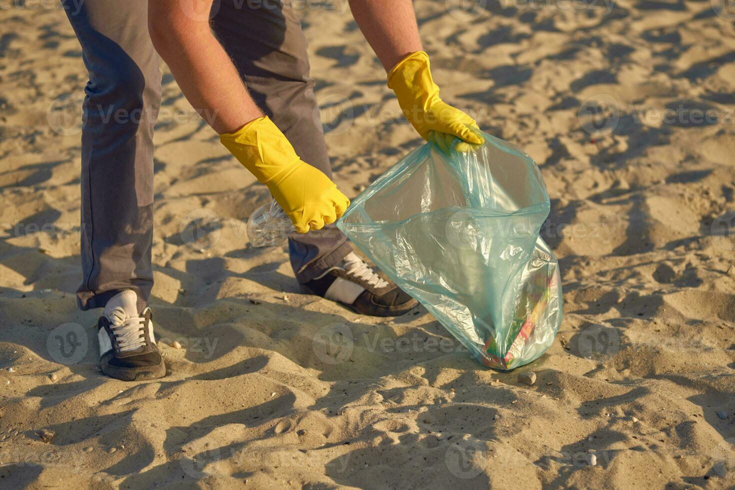 Young volunteer in yellow gloves is walking with garbage bag along a dirty beach of the river and cleaning up trash. People and ecology. Close-up. photo