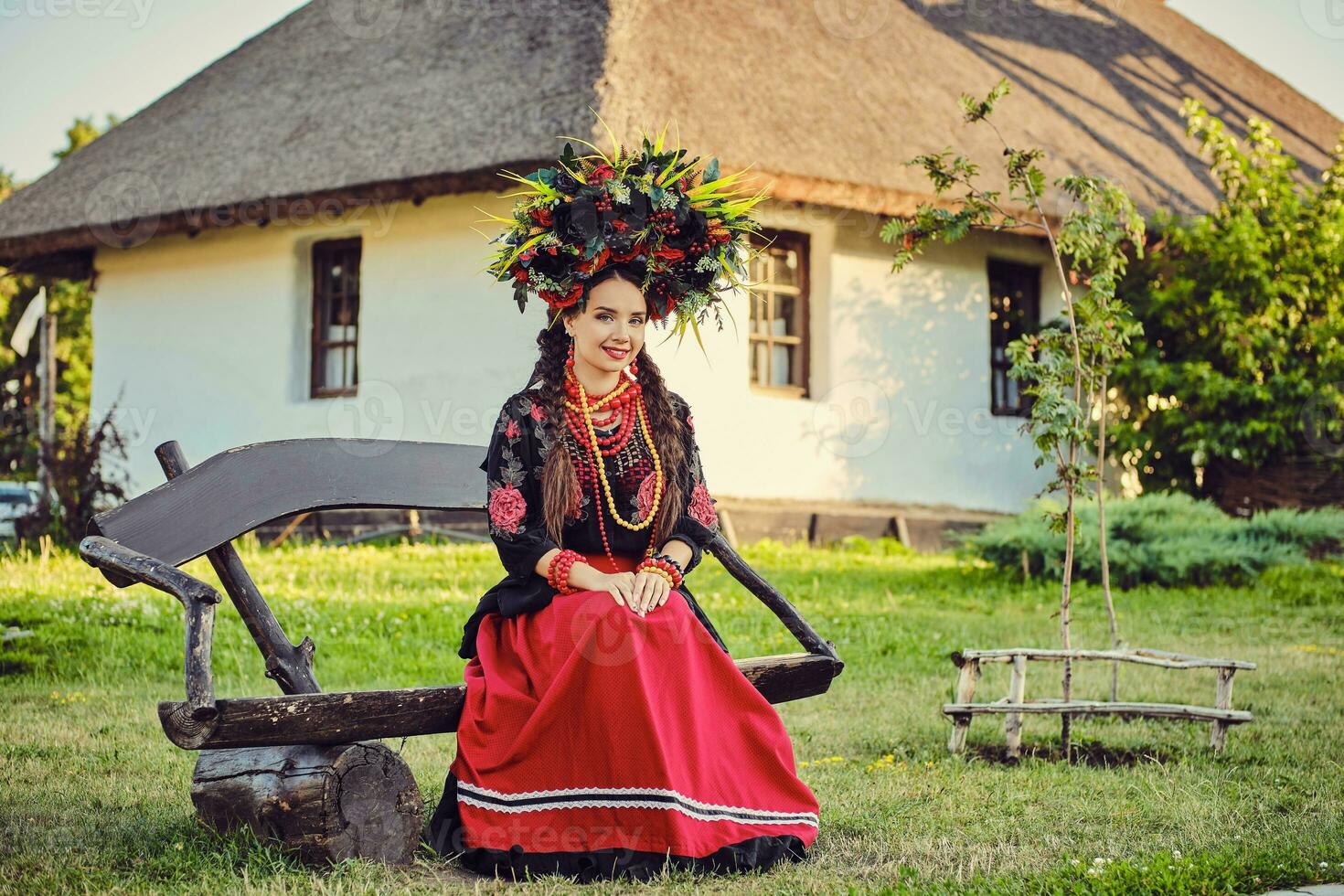 Brunette girl in a white ukrainian authentic national costume and a wreath of flowers is posing sitting on a wooden bench against a white hut. photo
