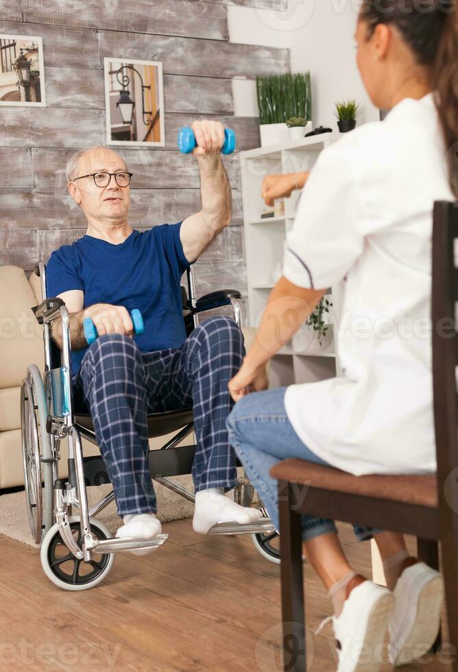 Grandfather in wheelchair doing sport assisted by nurse. Disabled handicapped old person with social worker in recovery support therapy physiotherapy healthcare system nursing retirement home photo