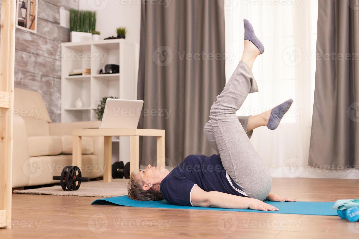 Old woman exercising in her apartment sitting on yoga mat. Old person pensioner online internet exercise training at home sport activity with dumbbell, resistance band, swiss ball at elderly retirement age photo