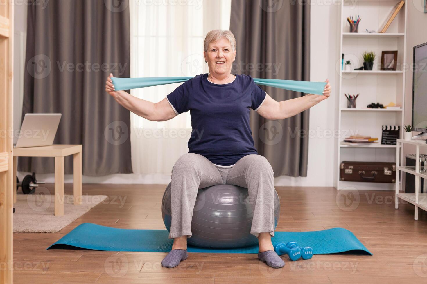 Aged woman enjoying exercise in her cozy apartment. Old person pensioner online internet exercise training at home sport activity with dumbbell, resistance band, swiss ball at elderly retirement age photo