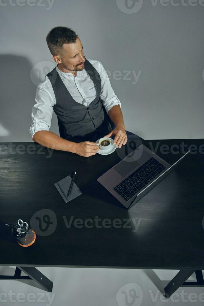 Bearded businessman dressed in classic suit is holding cup of coffee, sitting at table in office, looking away, gray background. Top view. photo