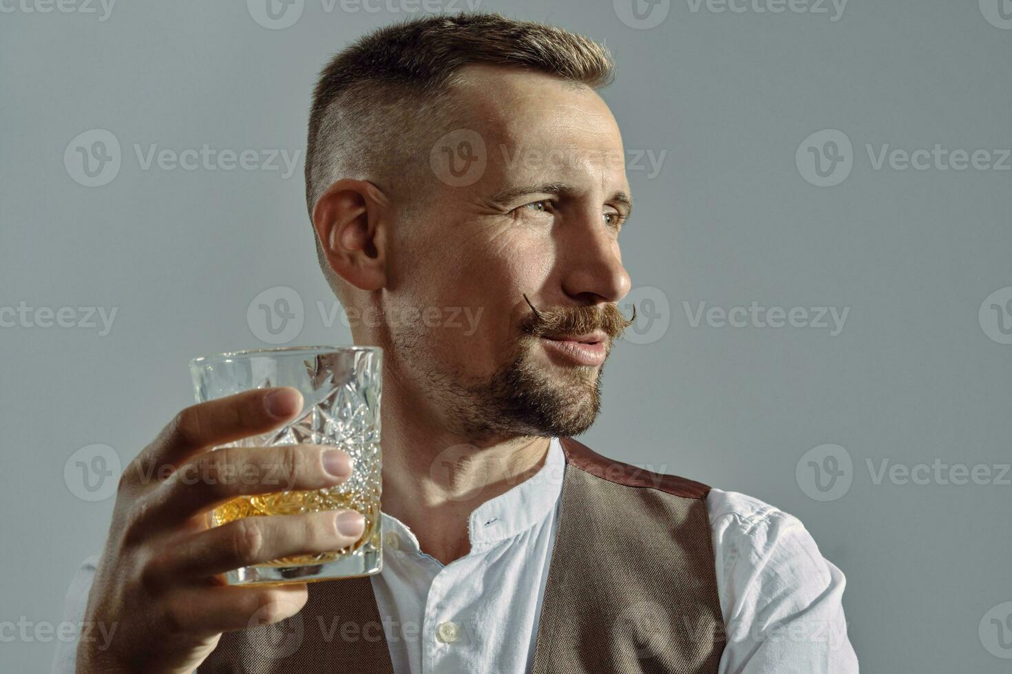 Man with stylish mustache, dressed in classic brown vest, white shirt is sitting at the table, enjoying whiskey. Grey background, close-up shot. photo