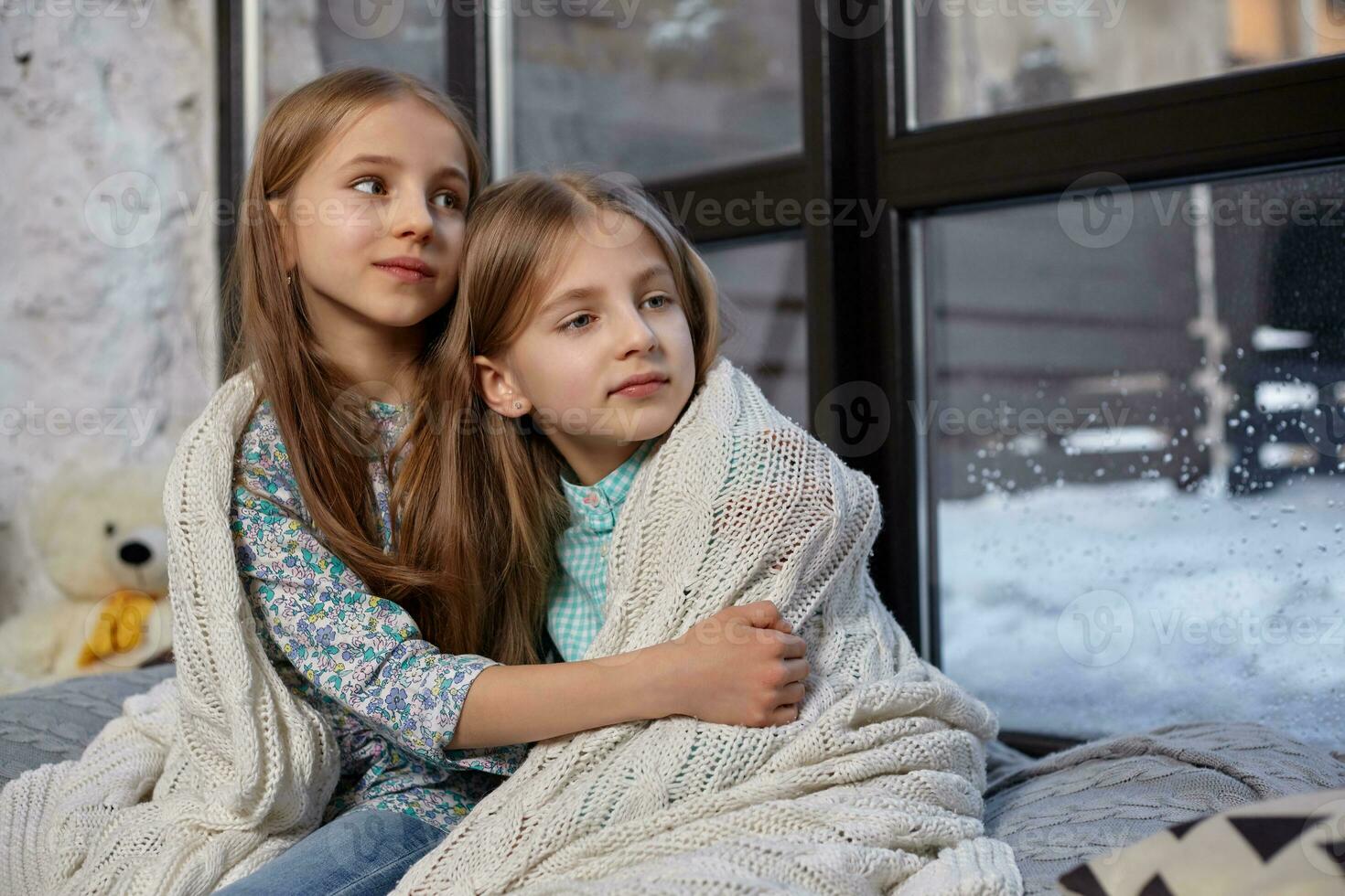 The image of two cute little sisters sitting on windowsill, covered by blanket, hugging each other photo