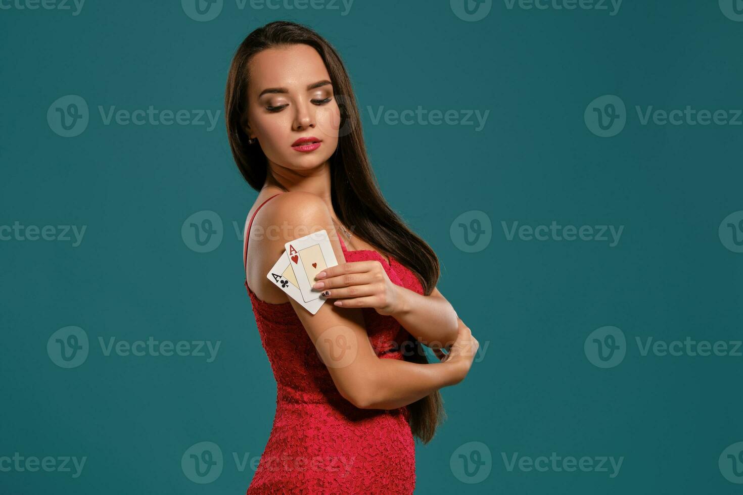 Brunette girl with a long hair, wearing a sexy red dress is posing holding two playing cards in her hand, blue background. photo