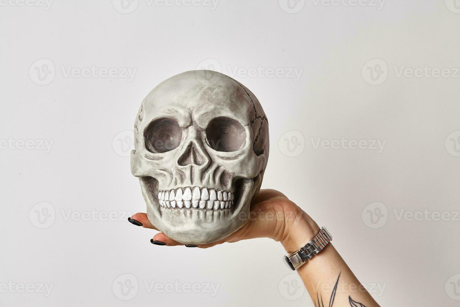 Tattooed hand of a woman in a black watch is holding a realistic model of a human skull with teeth isolated on white. Close-up shot. photo