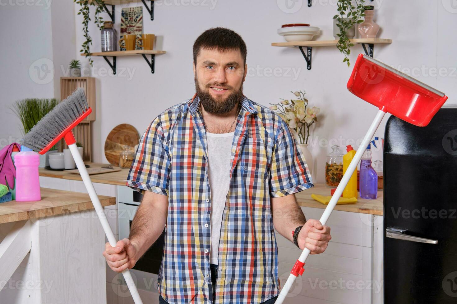 Young handsome bearded man in the kitchen, wearing checkered shirt, tries to deal with a dustpan and a broom. photo