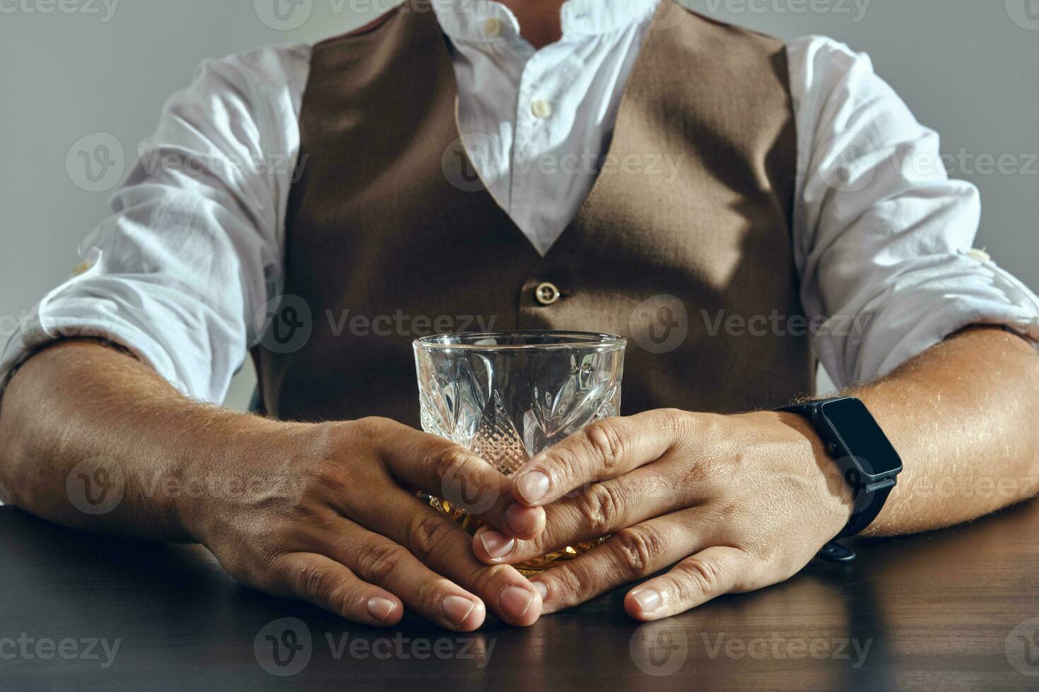 Hands of a man, dressed in a classic brown vest and white shirt is sitting at the table and enjoying whiskey. Grey background, close-up shot. photo