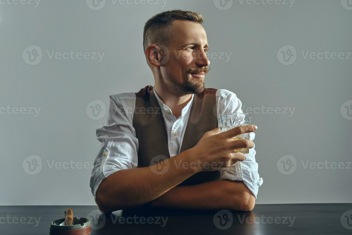 Man with stylish mustache, dressed in classic brown vest, white shirt is sitting at table, enjoying whiskey, cigar. Grey background, close-up shot. photo