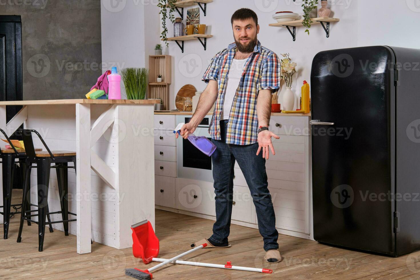 Young handsome bearded man in the kitchen, wearing checkered shirt, tries to deal with a dustpan and a broom. photo