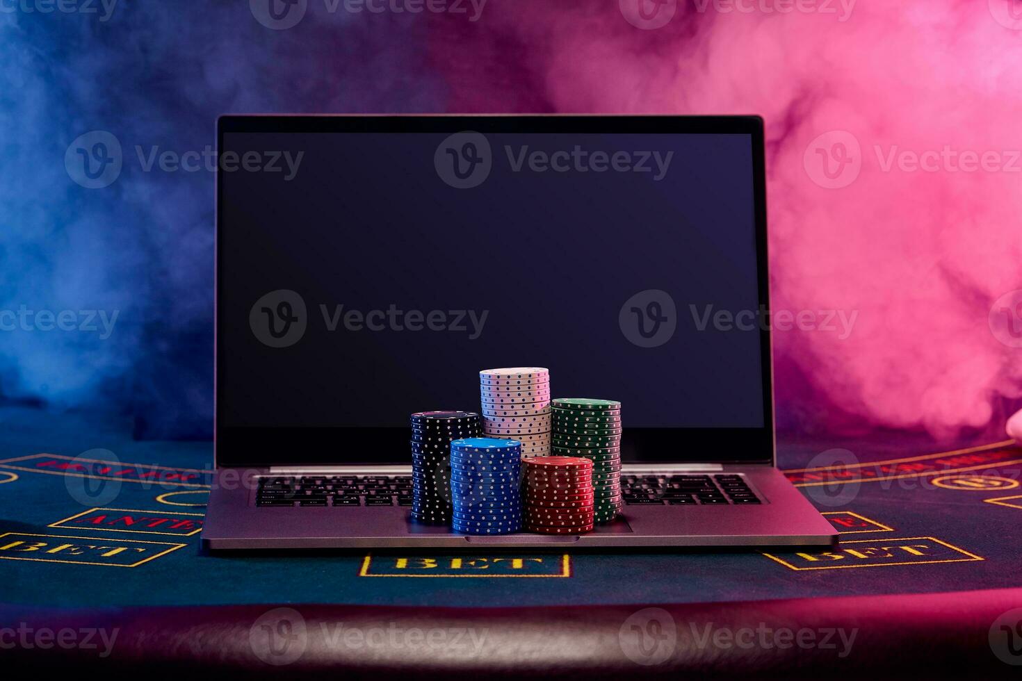 Chips piles are on laptop standing on blue cover of playing table. Black, smoke background. Gambling entertainment, poker, casino concept. Close-up. photo