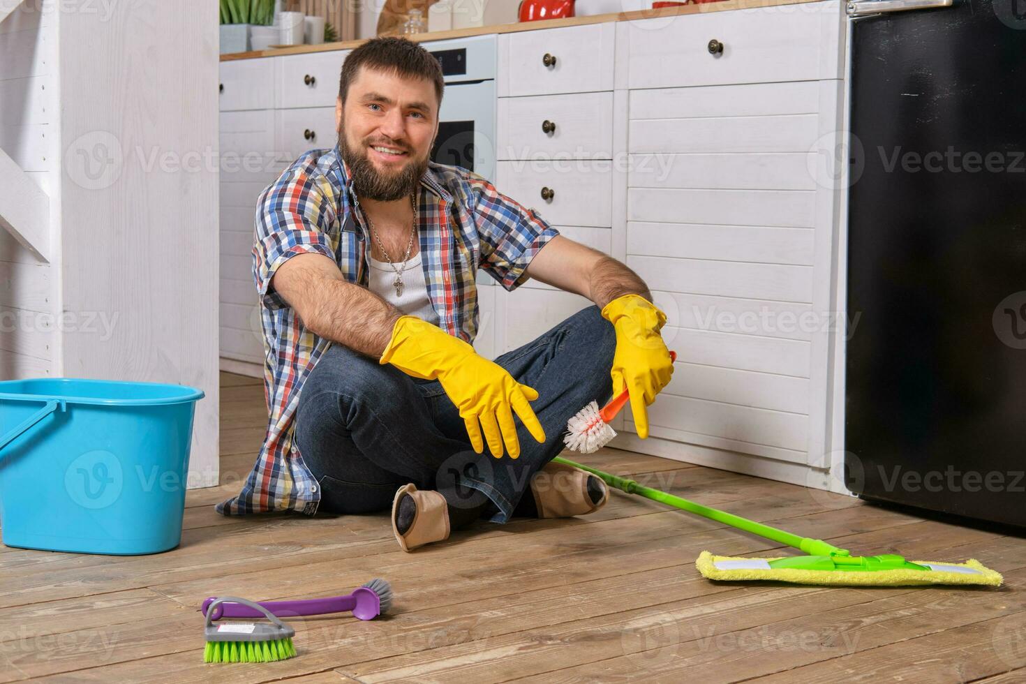 Caucasian young bearded man sits on the floor of his kitchen and tries to clean it using all of his detergents, rags, washcloths and brushes photo