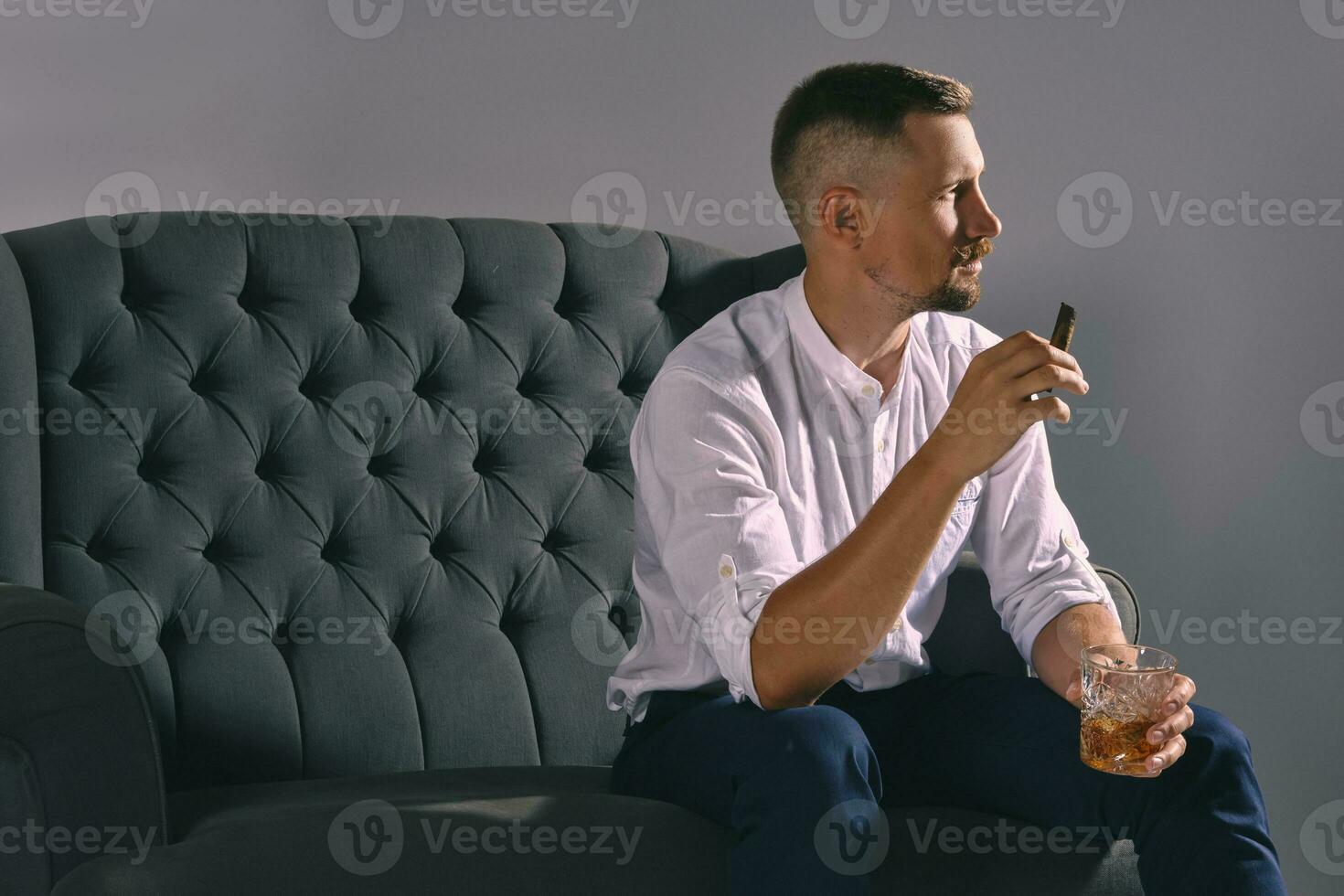Man with stylish mustache, dressed in white shirt, blue trousers is sitting on dark sofa, holding glass of whiskey, cigar. Grey background, close-up. photo