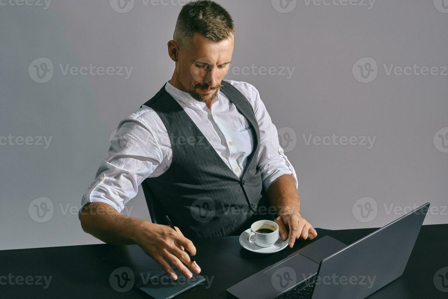 Bearded businessman with stylish mustache, dressed in a classic suit is working at laptop while sitting at table in office, gray background. photo