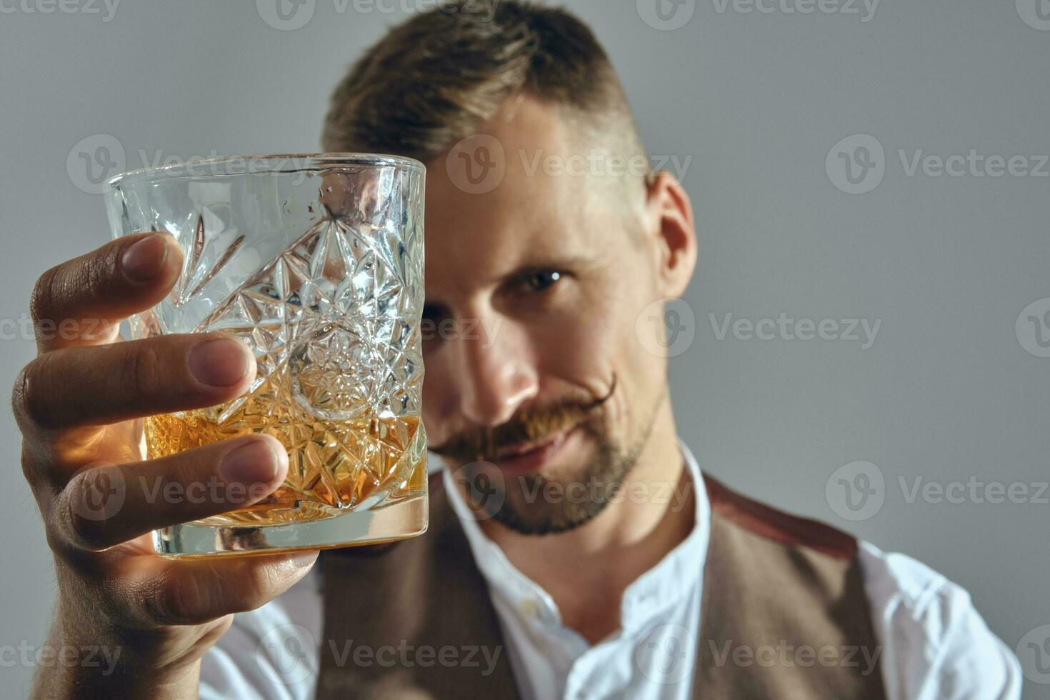 Man with stylish mustache, dressed in classic brown vest, white shirt is sitting at the table, enjoying whiskey. Grey background, close-up shot. photo