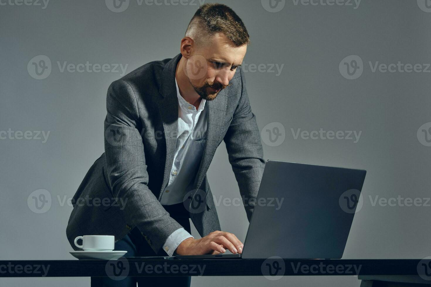 Bearded businessman with stylish mustache, dressed in a classic suit is working at laptop while standing at table in office, gray background. photo