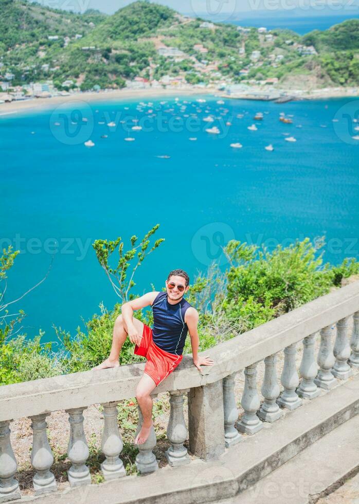 Portrait of tourist man in the view of the bay of San Juan del Sur. Happy tourist sitting in a viewpoint of a bay, Travel and tourism promotion concept. photo