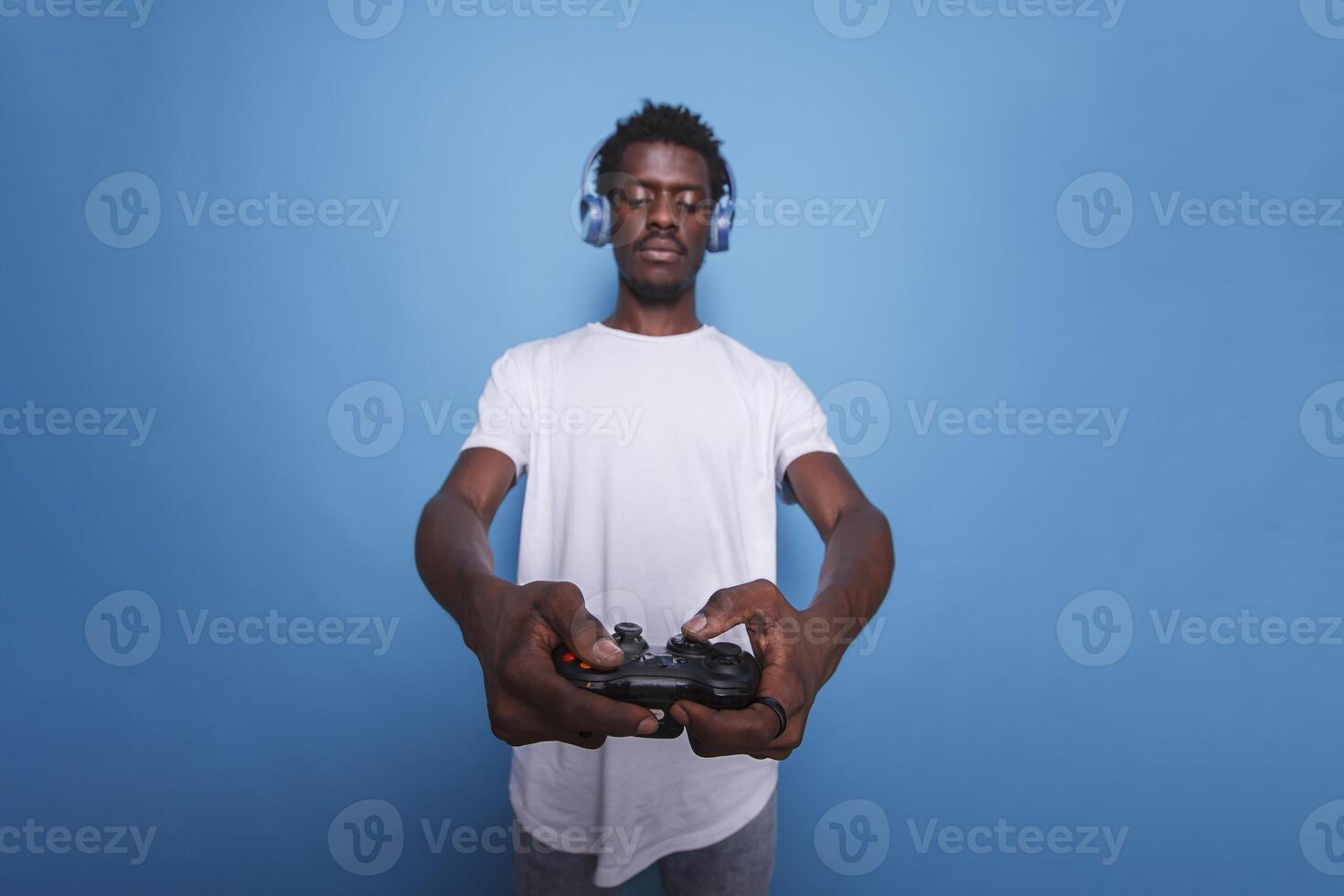 African American video gamer in studio using controller and headphones. Black man playing an internet game on a console with a joystick while standing over isolated blue background. photo