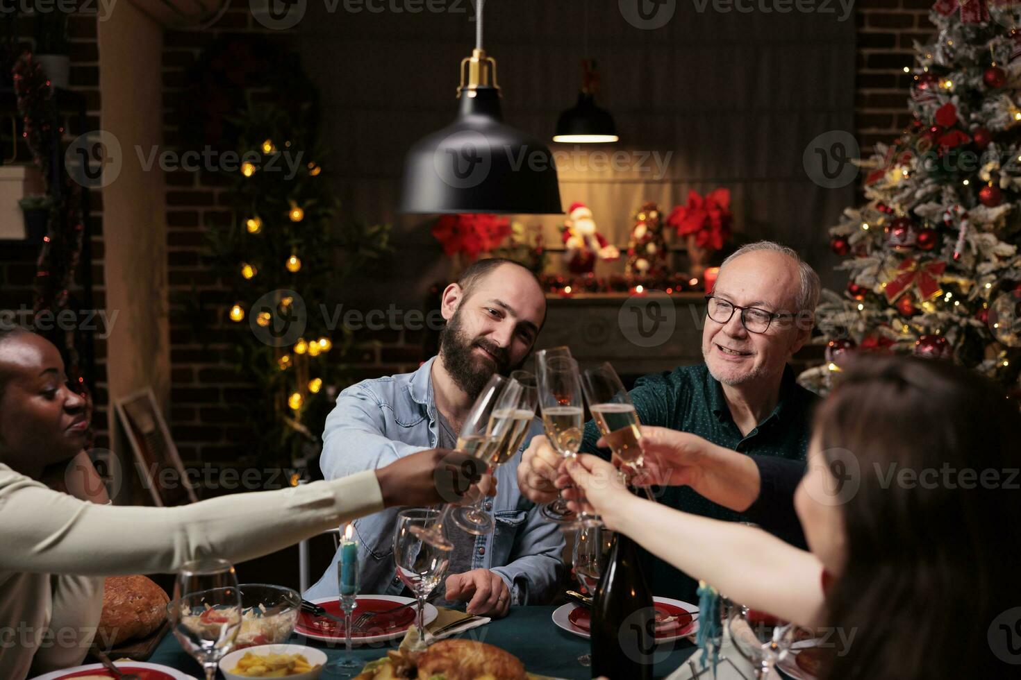 Diverse persons clinking alcohol glasses at christmas eve dinner, celebrating holiday tradition with wine and homemade meal. Friends and family saying cheers with drinks at table, cozy ornaments. photo