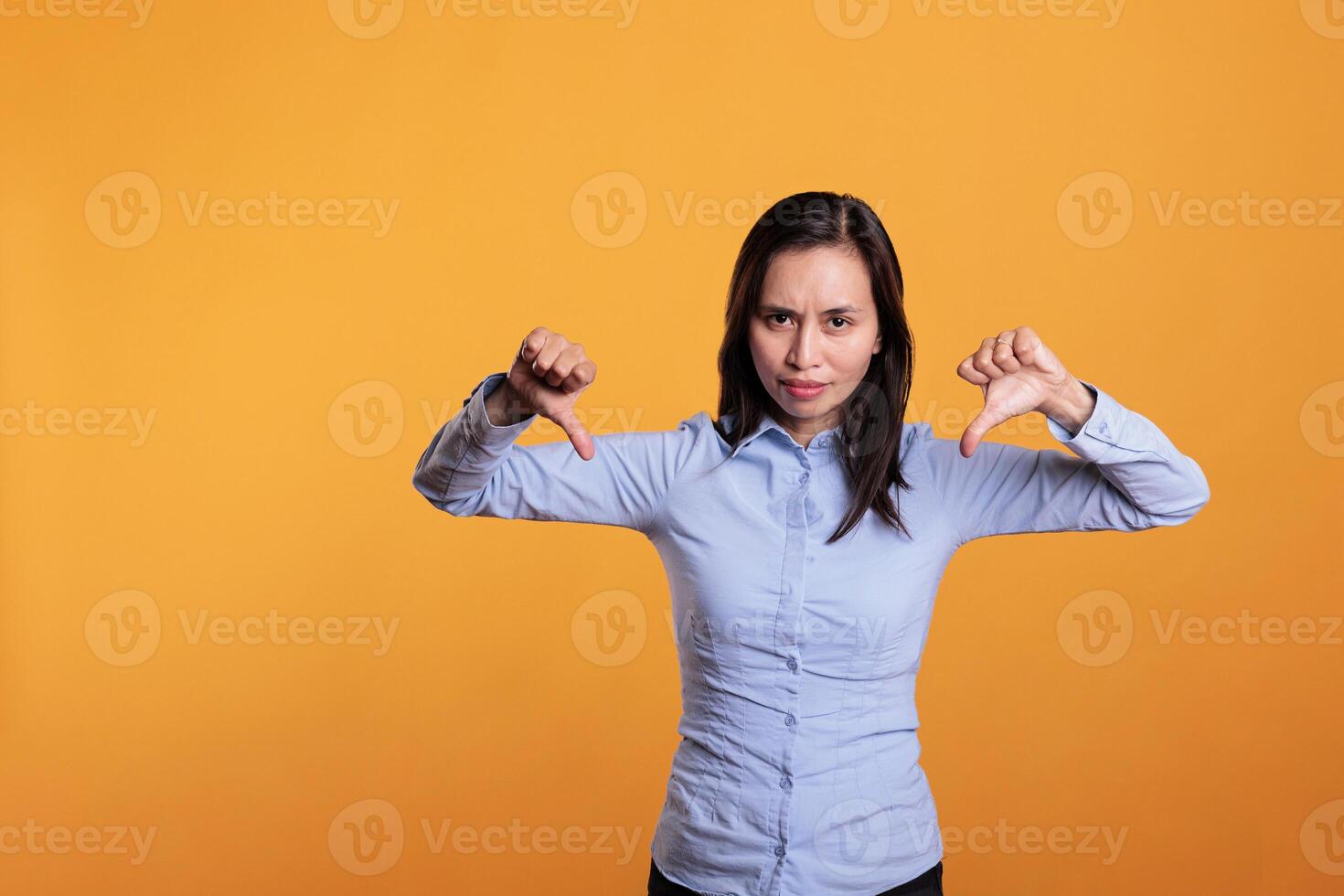 Unhappy filipino woman showing dislike gesture on camera in studio, gesturing disagreement sign with fingers. Displeased young adult expressing disapproval and disappointment sign. Dislike symbol photo