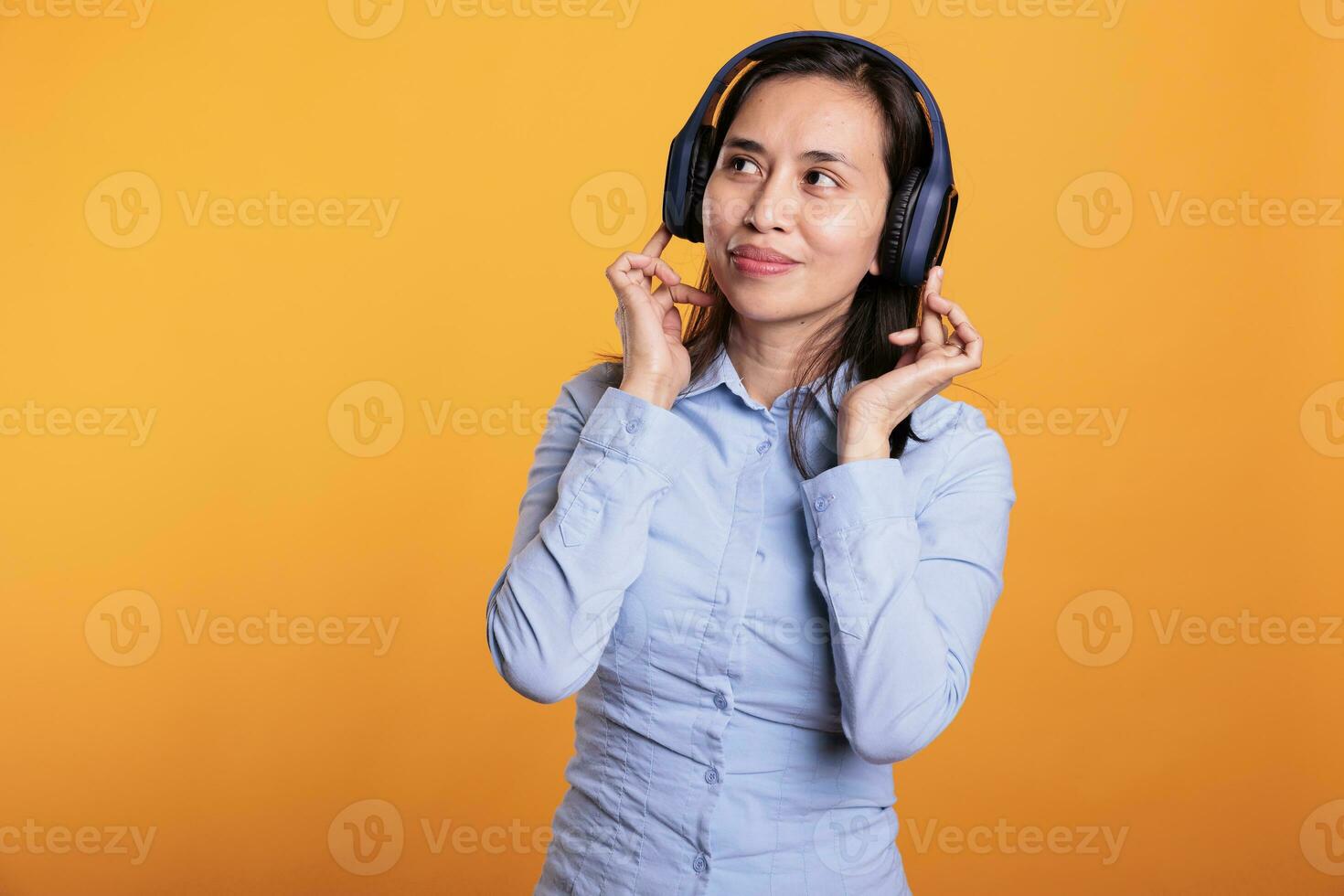 Cheerful woman with headset realaxing listening music, dancing over yellow background. Asian adult having fun in studio, showing dance moves during break time. Entertainment concept photo