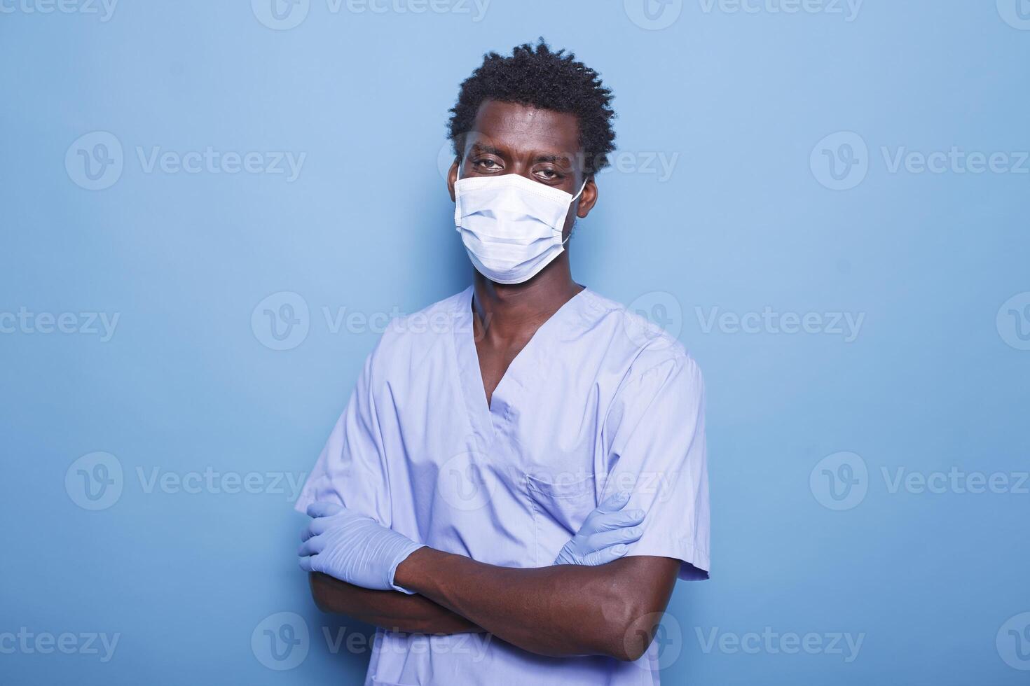 Portrait of black man wearing face mask and uniform standing in studio, working for healthcare. Medical assistant looking at camera, having protection against coronavirus. photo
