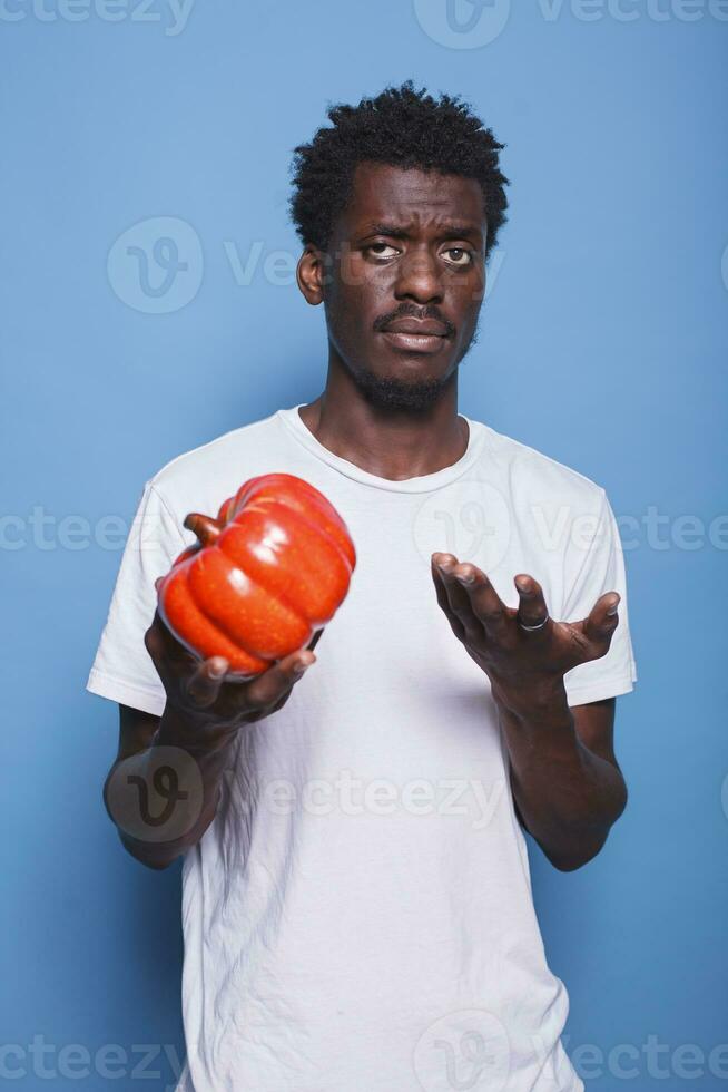 Black man holding red bell pepper in hand to cook meal for healthy diet. African american guy presenting natural vegetable for nutrition and vitamins, standing against isolated background. photo