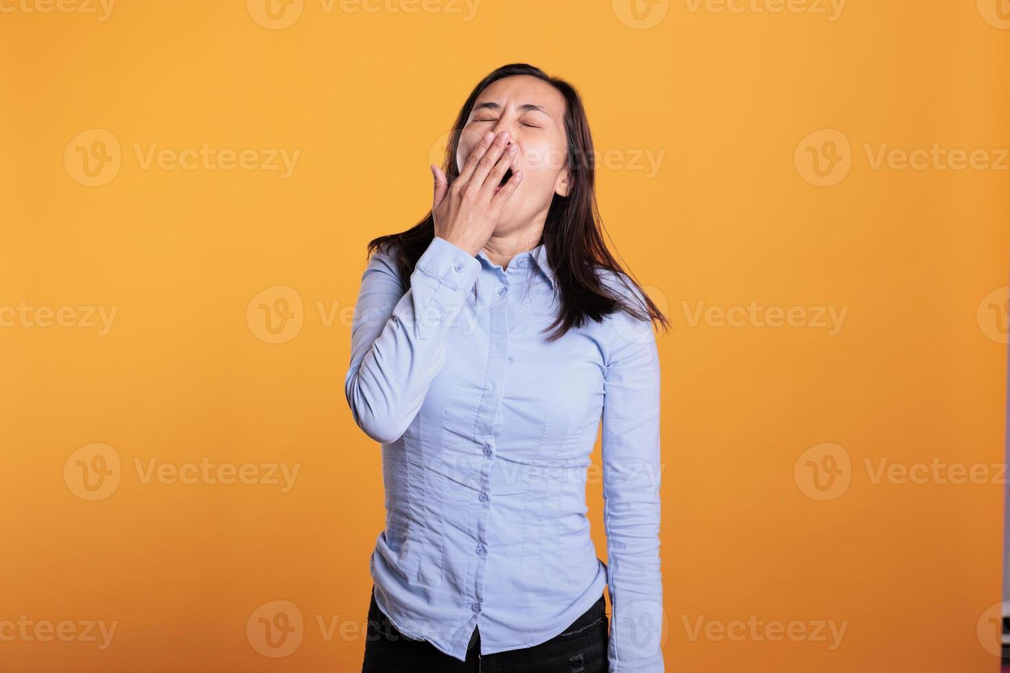 Portrait of sleepy filipino woman covering mouth with hand being tired while posing in studio standing over yellow background. Exhausted model yawning having lazy expression, working overtime photo