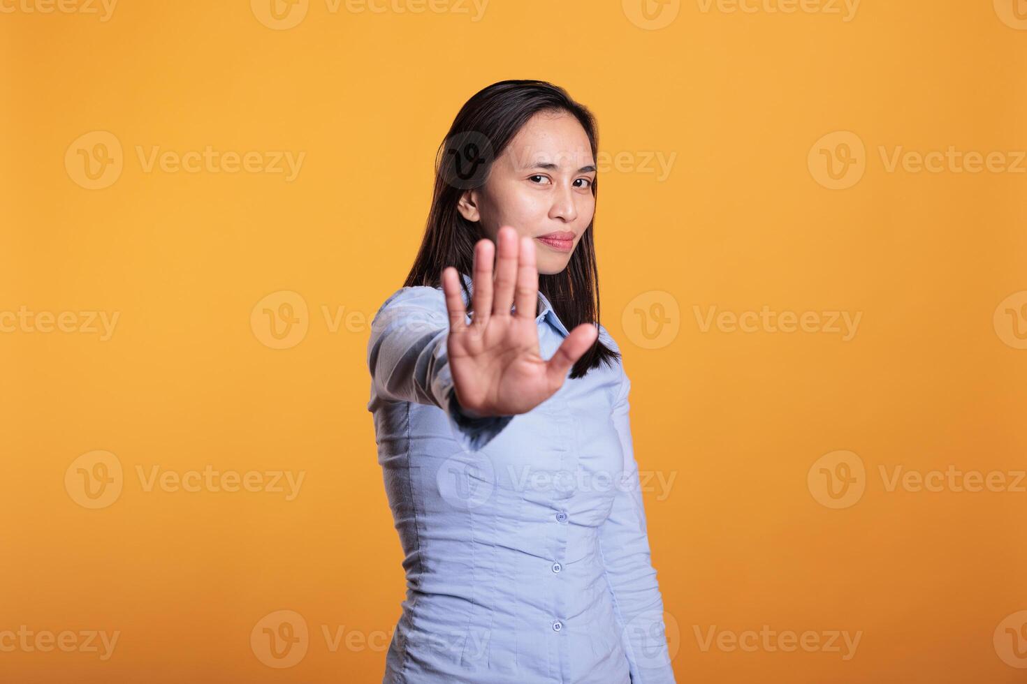 Filipino young adult doing no sign with palm on camera, expressing disapproval and denial. Cheerful woman showing rejection and refusal gesture standing in studio over yellow background photo