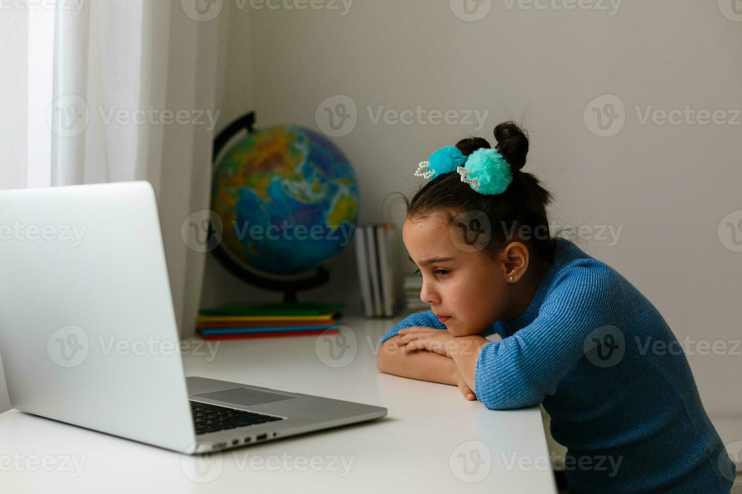sad little girl sitting in her room in front of open laptop pc, looking at screen while doing homework, studying online photo