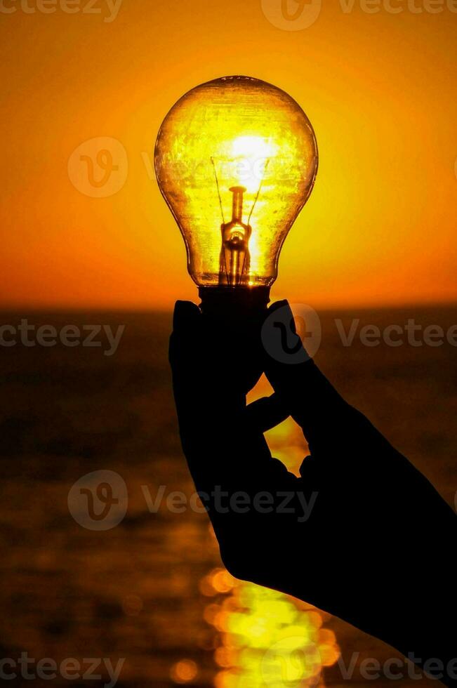 a person holding a light bulb over the ocean at sunset photo