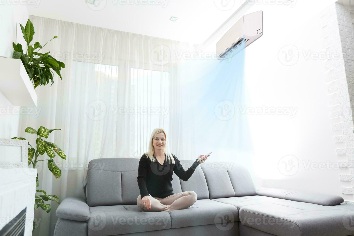 Young woman switching on air conditioner while sitting on sofa near white wall photo