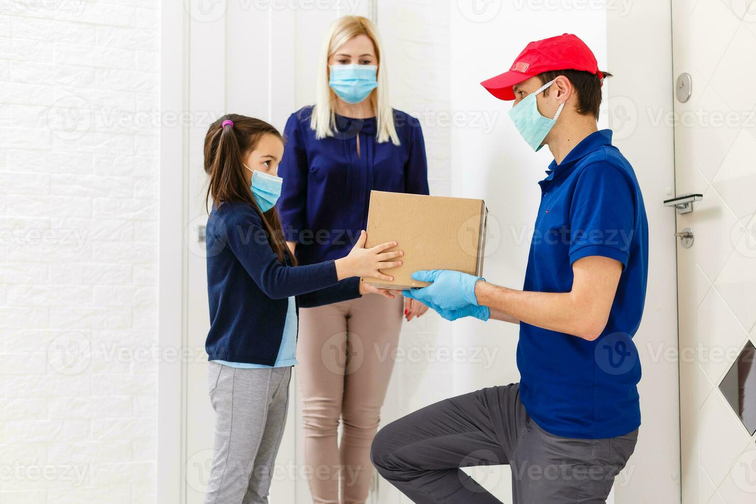 Young woman with little daughter wearing medical mask receiving orderd food from delivery man indoors. Prevention of virus spread photo