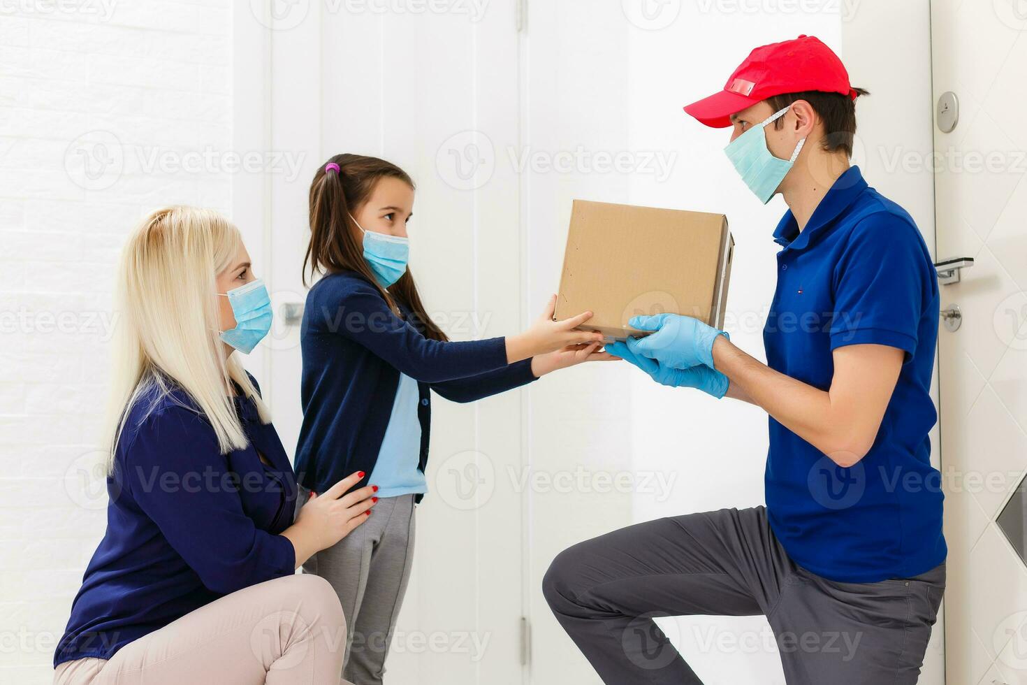 Young woman with little daughter wearing medical mask receiving orderd food from delivery man indoors. Prevention of virus spread photo