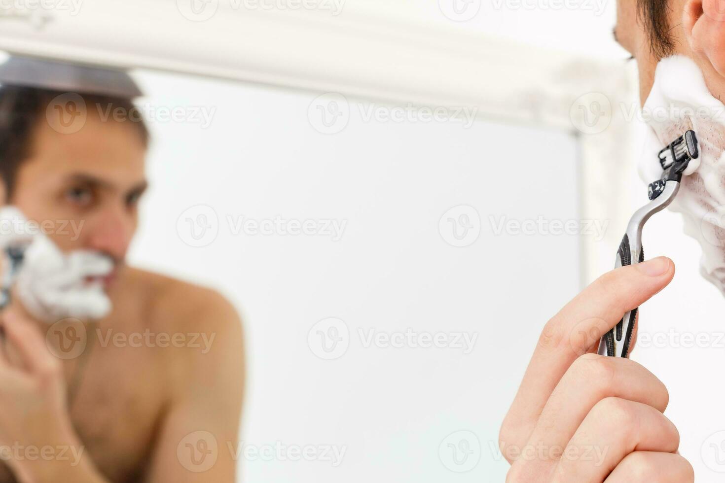 young man shaving in the bath. He is passing the razor for the beard while it looks at the mirror photo