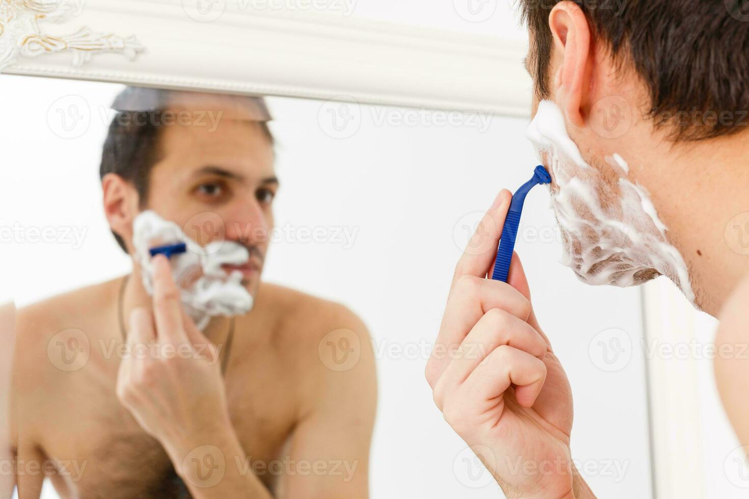 joven hombre afeitado en el baño. él es paso el maquinilla de afeitar para el barba mientras eso mira a el espejo foto