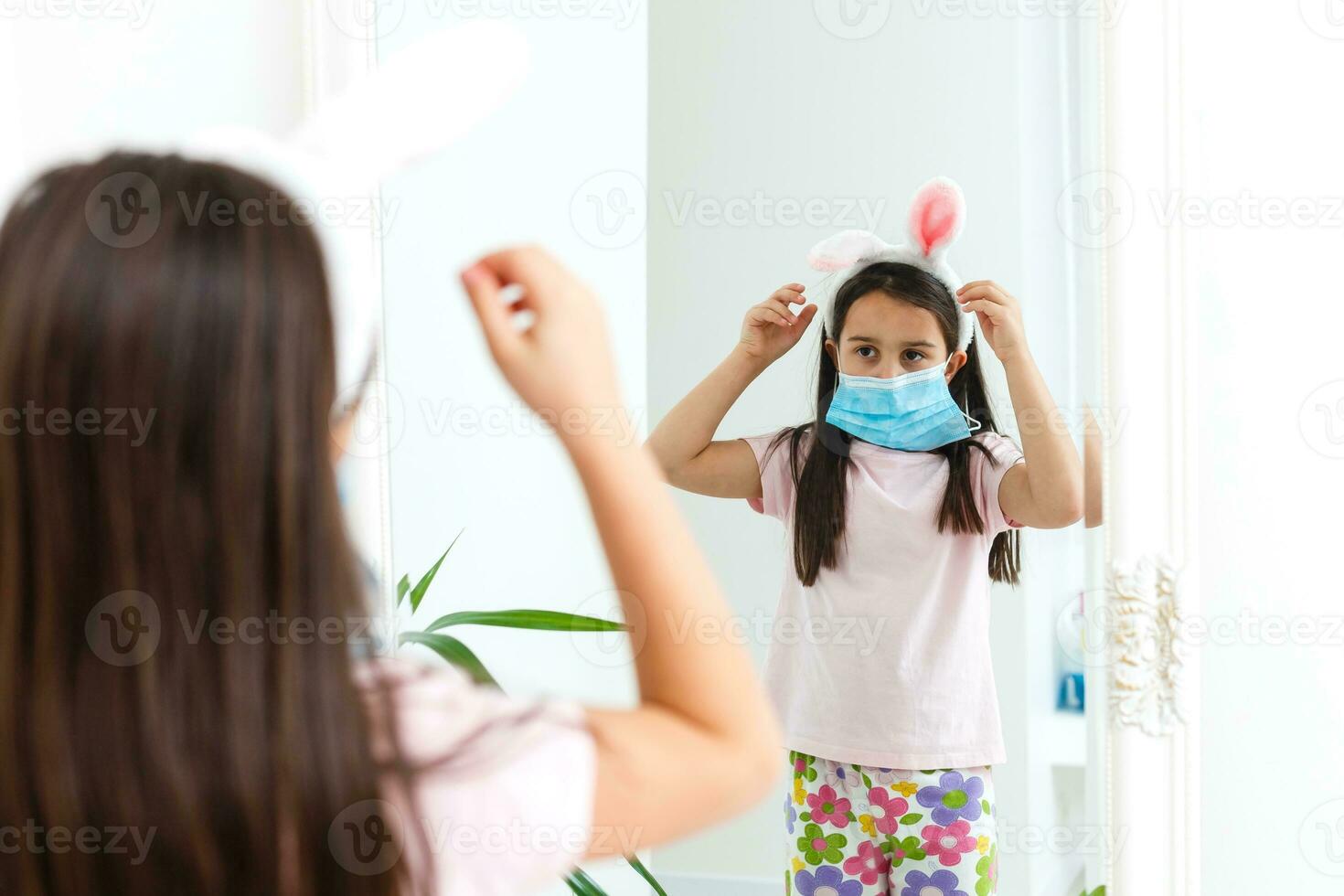 Little caucasian girl with bunny ears and medical mask on her face on a white background. Easter concept. Coronavirus protected photo