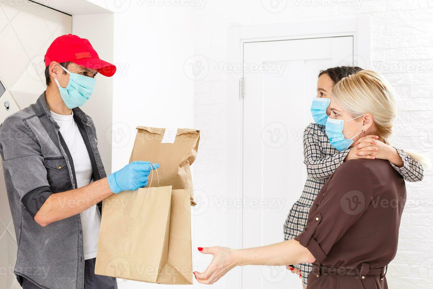 Young woman wearing medical mask receiving orderd food from the restaurant from delivery man indoors. Prevention of virus spread photo
