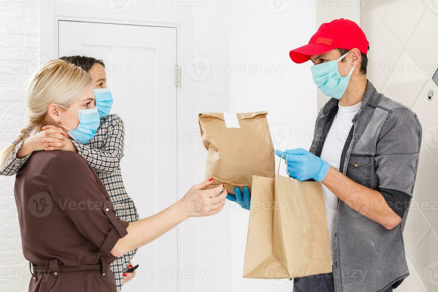 Young woman wearing medical mask receiving orderd food from the restaurant from delivery man indoors. Prevention of virus spread photo