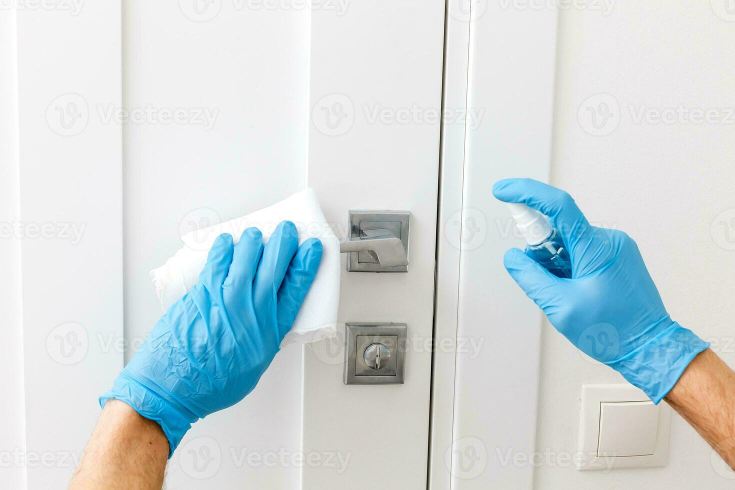 Hands in protective gloves - one holds the sprayer with an antiseptic, the other - wipes the door handle with a cloth moistened with a disinfectant solution photo