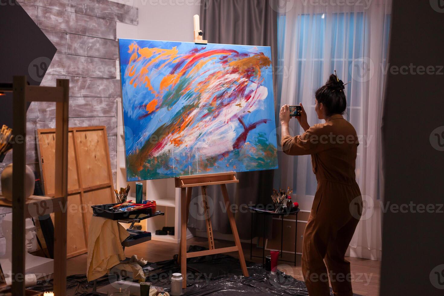 Artist using phone to take a photo of her masterpiece painting in art workshop. Modern artwork paint on canvas, creative, contemporary and successful fine art artist drawing masterpiece