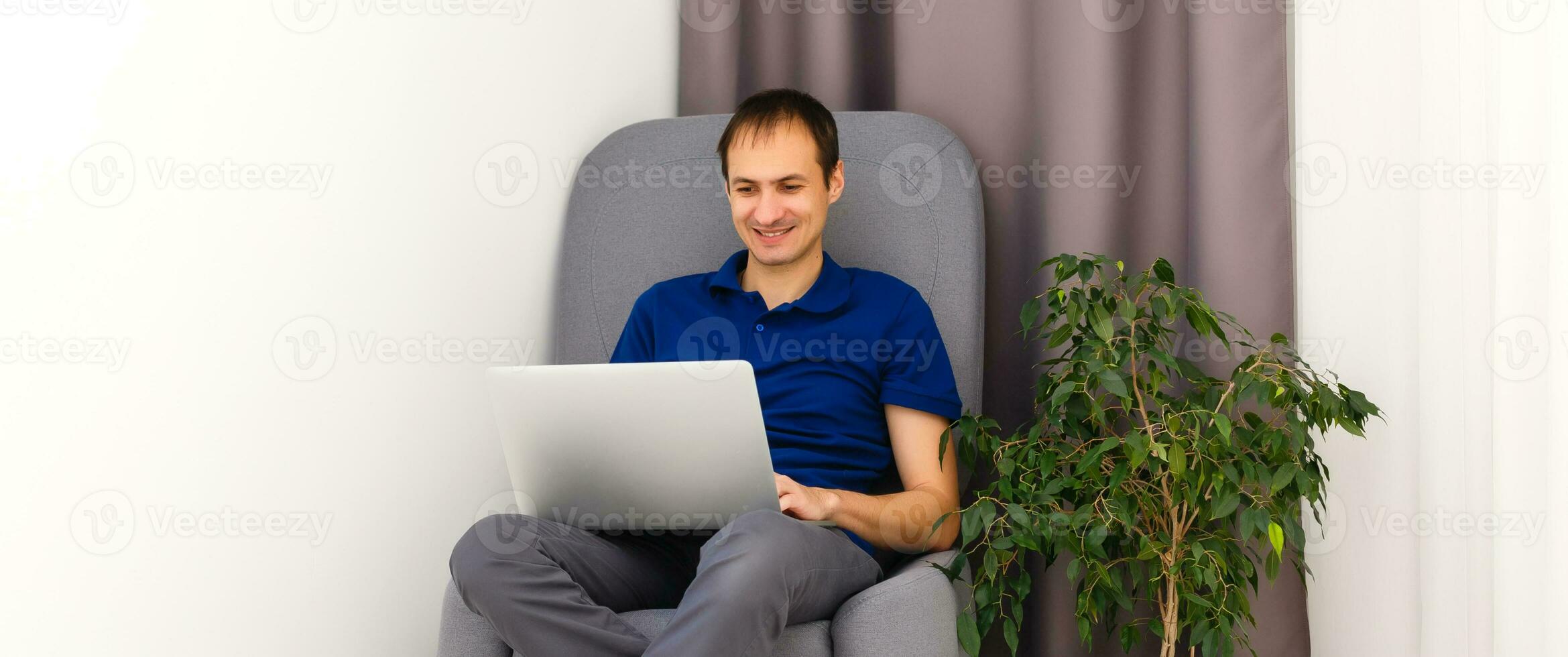 Happy young man in t-shirt sitting on sofa at home, working on laptop computer, smiling. photo